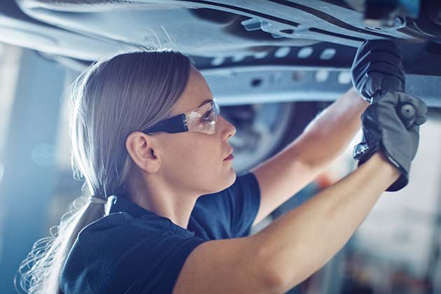 A female automotive technician wearing safety glasses and gloves works underneath a vehicle in a garage
